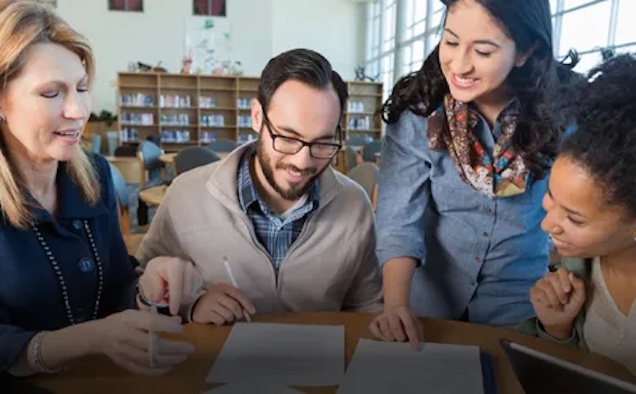 A group of four diverse teachers look down and smile at collaborative work.